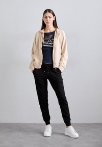 Beżowa bluza z zamkiem na przodzie noszona na granatowy t-shirt z grafiką, w zestawie z czarnymi dresami i białymi sneakersami. Gładki materiał, luźny krój, pas ze sznurkiem.
