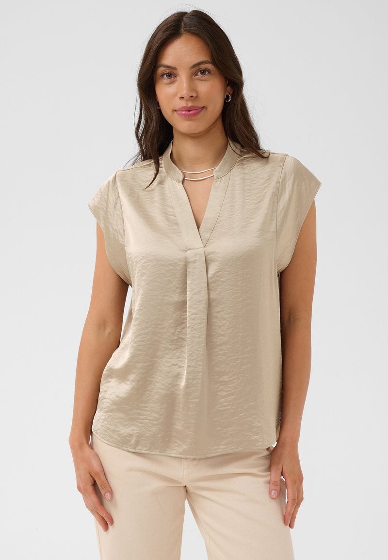 Femme aux longs cheveux foncés portant un chemisier en satin beige à manches courtes et un pantalon de couleur claire, debout devant un fond uni.