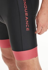 Svarta träningsshorts med en rosa midja. Har "ENDURANCE" tryckt i fet röd text på sidan. Slät, stretchig tyg.