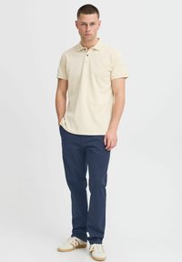 Svetlo bež polo majica s kratkimi rokavi in subtilnim logotipom, kombinirana z mornarsko modrimi hlačami in bež teniskami. Gladka tekstura tkanine.