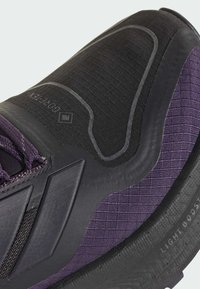 Chaussure de sport noire et violette avec un tissu texturé, une semelle en caoutchouc et des motifs de design distinctifs. Comprend un marquage visible et des accents matériels.