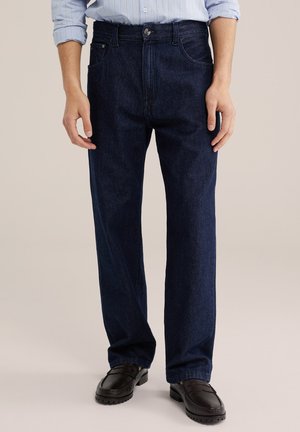 Mand iført mørkeblå jeans med lige ben, lyseblå stribet opsmøget skjorte og sorte loafers, stående mod en neutral baggrund.