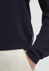 Pulover tricotat de culoare bleumarin, cu manșete și tiv cu nervuri, având o textură interesantă și o etichetă mică cu logo pe lateral.