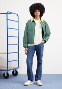 Green quilted jacket with a beige collar, beige graphic t-shirt, blue jeans, and white shoes with a chunky sole. Blue cart in background.