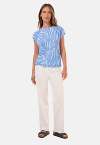Blusa azul de manga corta con patrones de hojas blancas, combinada con pantalones blancos y sandalias oscuras. La blusa tiene un escote relajado y una tela texturizada.