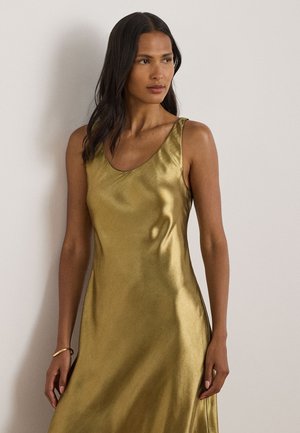 Femme aux longs cheveux foncés portant une robe en satin dorée sans manches, debout contre un mur uni de couleur claire.