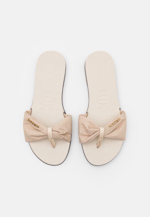 Par beige flade sandaler med stofsløjfe-remme og tåadskillere, teksturerede fodsenge og mærkede guldfarvede mærker på hvid baggrund.