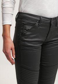 Pantalones de imitación cuero negros con detalles acanalados y herrajes plateados. Presenta una etiqueta de G-Star en el bolsillo frontal. Textura suave.