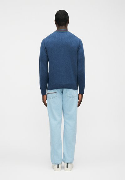 Homme portant un pull bleu avec un col rayé, un jean bleu clair et des baskets blanches, debout face à un fond blanc uni.