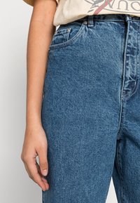 Jeans de ganga azuis com cintura alta, apresentando um clássico design de cinco bolsos e detalhes de costura subtil, mostrados da cintura para baixo com uma mão.