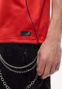 Κόκκινο αθλητικό πουκάμισο με μαύρο περίγραμμα, φορεμένο με μαύρο παντελόνι με ασημένια αλυσίδα να κρέμεται από την τσέπη κοντά σε χαλαρό χέρι.