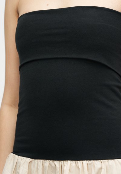 Γυναίκα που φοράει μαύρο τοπ χωρίς τιράντες και ανοιχτό μπεζ φούστα, μπροστά σε απλό φόντο, εστιάζοντας στο πάνω μέρος του κορμού.