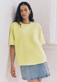 Maglietta oversize verde lime con collo rotondo e tasca, abbinata a una minigonna in denim chiaro. Tessuto liscio e vestibilità casual.