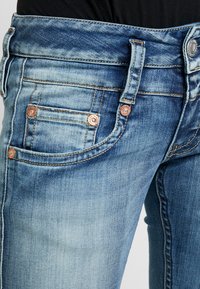 Blå denimjeans med en lätt blekt finish, utrustade med fem fickor, kopparnitar och kontrasterande sömmar längs sömmarna.