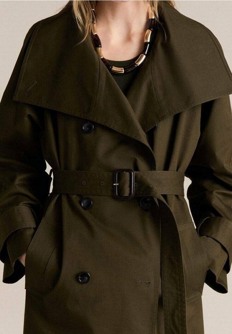Gros plan d'une personne portant un trench-coat vert foncé ceinturé avec de grands revers et des boutons noirs, accessoirisé d'un collier épais noir et or.