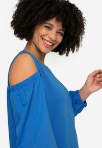 Blå avskuret blus med långa puffärmar och elastiska manschetter, tillverkad av ett mjukt tyg och har en strukturerad yta.