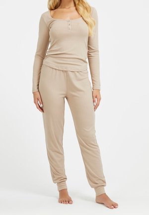 Woman wearing beige ribbed long-sleeve top with buttoned neckline and matching ribbed jogger pants, standing barefoot on white background.