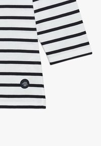 T-shirt à manches longues avec des rayures horizontales blanches et marine, doté d'un patch logo rond sur le côté gauche. Tissu en coton doux.