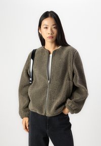 Oliivivihreä fleece-takki, jossa on etuvetoketju, pyöreä kaula-aukko ja joustavat hihat, yhdistetty tummaan farkkuihin ja mustaan hihnaan.