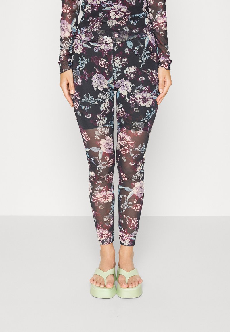 Leggings com estampado floral em tecido preto translúcido, apresentando flores em roxo, azul e rosa, complementadas por sandálias de plataforma verde-claro.