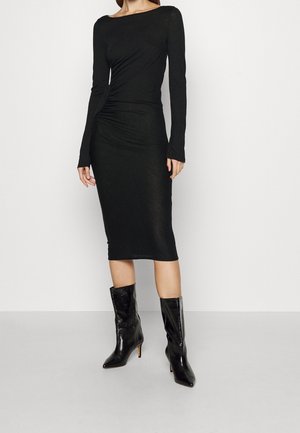 Femme portant une robe midi noire ajustée à manches longues avec fronces sur les côtés, associée à des bottes noires à talons mi-mollet et bouts pointus.