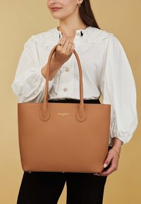 Sac fourre-tout en cuir tan avec deux longues anses, présentant une surface texturée et un logo en relief. Lignes épurées et design minimaliste.