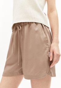 Light brown shorts made of smooth fabric with an elastic waistband and adjustable drawstring. Side pockets and stitched hem detail visible.