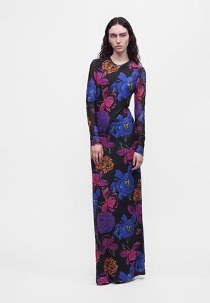Un mannequin se tient devant un fond blanc, vêtu d’une longue robe noire ornée de motifs floraux bleu vif, rose et orange.