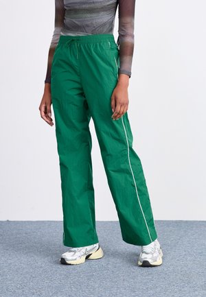 Person wearing loose green track pants with white side stripes and silver athletic shoes, standing on gray carpeted floor.