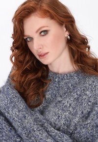 Grauer und blauer Strickpullover mit einem Texturmuster; das Model hat lange, lockige rote Haare, die die Gesichtszüge mit minimalem Make-up betonen.