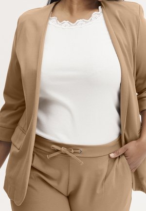 Femme portant un blazer beige et un pantalon à cordon à la taille, haut blanc côtelé avec bordure en dentelle, main dans la poche, torse visible.