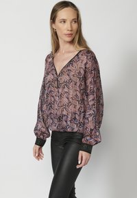 Durchsichtige Bluse mit langen Ärmeln und tiefem V-Ausschnitt, die ein Paisleymuster in Lila- und Brauntönen aufweist. Schwarze strukturierte Manschetten runden das Design ab.