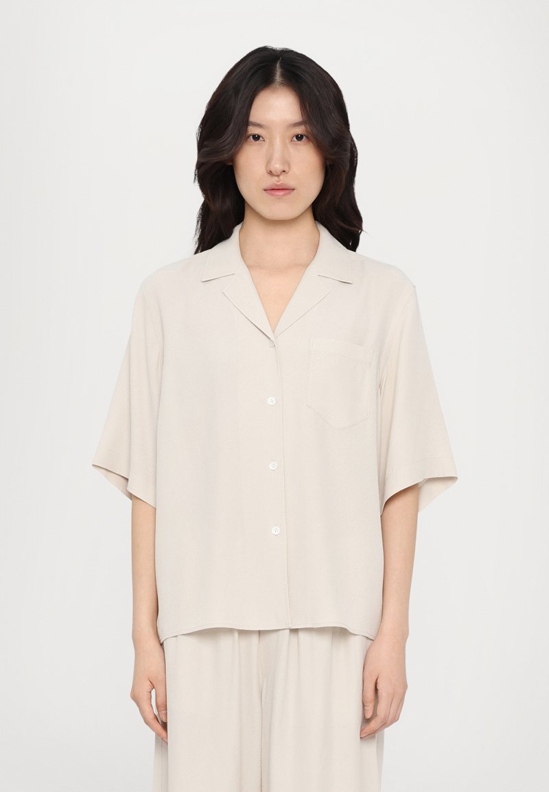 Lys beige kortærmet skjorte i et blødt materiale, med krave, knaplukning og en frontlomme. Enkelt, afslappet design.