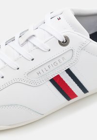 Białe sneakersy wykonane z materiału syntetycznego, z teksturowanym wzorem, sznurówkami i logo. Udekorowane czerwonymi i granatowymi paskami.