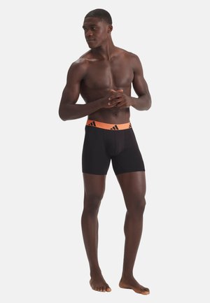 Athletischer Mann steht barfuß, trägt schwarze Boxer-Shorts mit einem orangenem Bund, der schwarze Adidas-Logos zeigt.