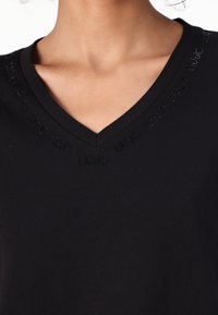 T-shirt nera a V realizzata in morbido cotone, con sottile marchio in rilievo nero attorno al colletto, che offre una trama liscia.