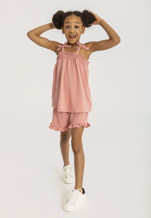 Roze mouwloze top met gerimpelde details en strikjes op de schouderbanden, gecombineerd met bijpassende shorts met ruches aan de zoom, gedragen met witte sneakers.