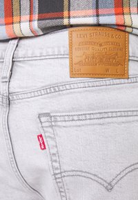 Zadní kapsa světle šedých džínsů Levi's s hnědou koženou značkovou nášivkou a červenou nášivkou Levi's, spárovaná s kostkovanou košilí.