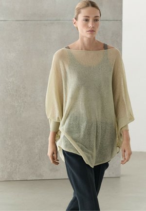 Ung kvinde iført en gennemsigtig beige oversize sweater over en grå top og mørke bukser, stående indendørs mod en lysegrå væg.