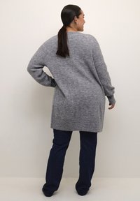 Langer grauer Kabelstrickpullover mit geripptem Saum und Bündchen, kombiniert mit dunklen Jeans. Die Textur ist weich und hat ein lässiges Design.