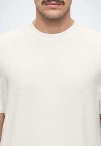 Homme portant un pull blanc texturé à manches courtes avec un col rond et une vue partielle de son visage et de sa moustache.