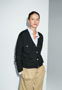 Cardigan lavorato a maglia nero con bottoni dorati e due tasche frontali, indossato sopra una camicia azzurra chiara e abbinato a pantaloni beige. Texture liscia.