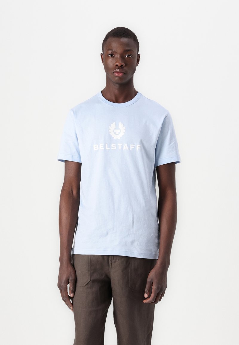 Gaiši zils kokvilnas T-krekls ar īsām piedurknēm, priekšā ar baltu 'BELSTAFF' logotipu un simbolu. Valkāts ar brūnām biksēm.