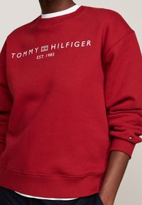 Rode sweatshirt met witte "TOMMY HILFIGER" logo en "EST. 1985" tekst. Heeft ribgebreide boorden en onderrand, met een klein logo accent op de mouw.