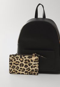 Sac à dos en tissu noir avec un sommet arrondi et une fermeture éclair. Accompagné d'un portefeuille en imprimé léopard avec une fermeture éclair et des accessoires dorés.