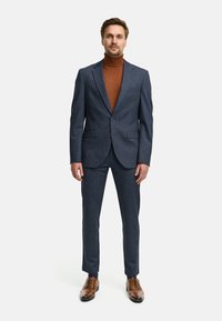 Marineblauw gestructureerd pak met een enkelgeknoopt colbert en twee voorzakken, gecombineerd met een bruine turtleneck en bruine dresschoenen.