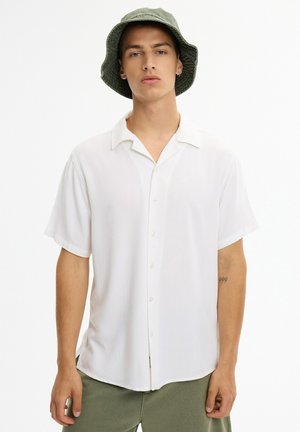 Joven con camisa blanca de manga corta con botones y sombrero verde tipo pescador, de pie frente a un fondo blanco liso.