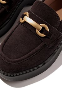 Donkerbruine suede loafers met een ronde neus, voorzien van een goudkleurige metalen kettingaccent en een zwarte gestructureerde rubberen zool.