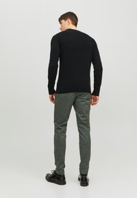 Maglione nero a coste e pantaloni verdi con texture. L'individuo indossa scarpe nere, visto da dietro su uno sfondo chiaro e neutro.