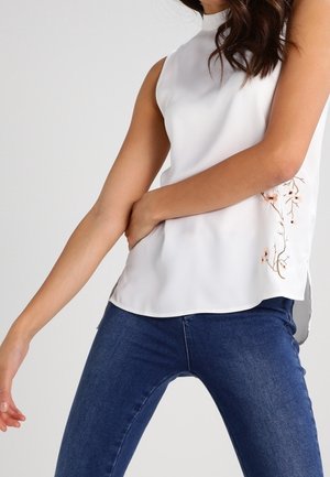 Woman wearing sleeveless white blouse with floral embroidery on the side and blue jeans, posing with one arm across her waist.